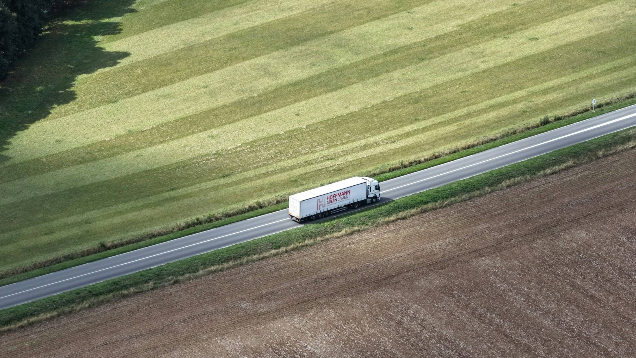 A semi truck driving down an empty highway between green fields, seen from above
