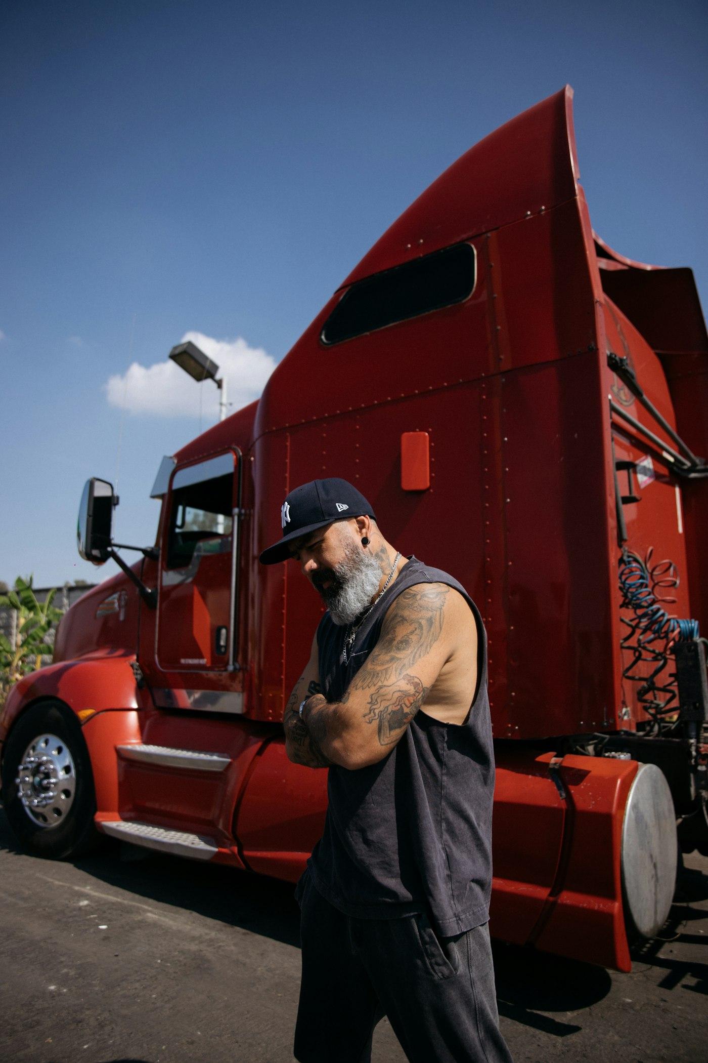 A bearded owner-operator in a cap standing in front of his red semi truck